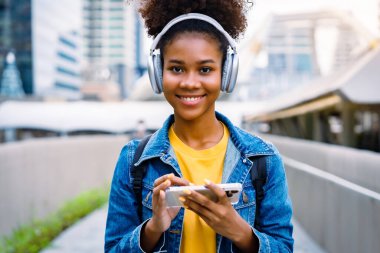 Cheerful black student girl wearing headset and using mobile smartphone, Walking at college building outdoor, Smiling and looking at camera. Modern education