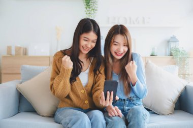 Evdeki oturma odasında gülümseyen ve selfie çeken iki Asyalı genç kadın. Video çağrısı, Toplantı konferansı