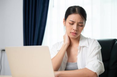 Ofis sendromu konsepti. Bilgisayarda uzun süre çalıştıktan sonra boyun ve omuz ağrısı çeken Asyalı genç bir kadın. Kaslarını gevşetmek için esniyor.