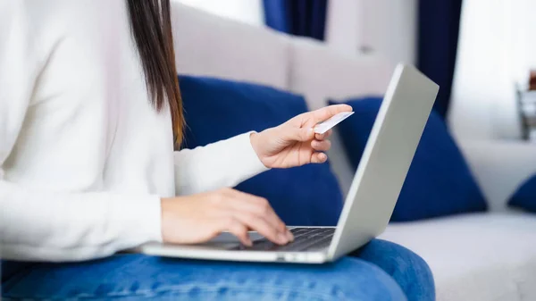 年轻的亚洲女人坐在客厅的沙发上 用电脑笔记本电脑上的银行卡通过互联网进行网上银行支付 用信用卡在笔记本上购物 — 图库照片