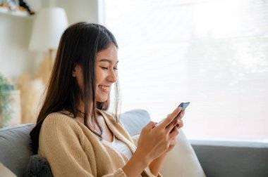 Happy young asian woman relax on comfortable couch at home texting messaging on smartphone, smiling girl use cellphone chatting, browse wireless internet on gadget, shopping online from home