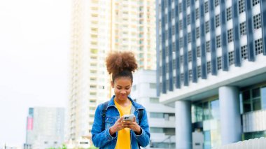 Zeki telefon kullanan ve şehirde yürürken gülümseyen genç siyahi bir kadın. Mutlu Afrikalı öğrenci, cep telefonuyla bir binanın önünden geçen genç bayan.