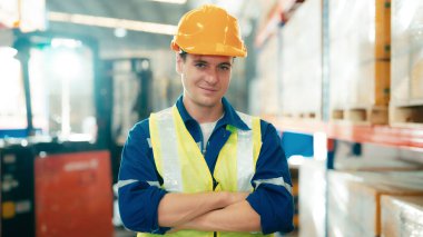 Handsome man and happy professional worker wearing safety vest and hard hat, Smiling to camera. Big warehouse with shelves full of stock