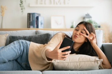 Happy young asian woman relax on comfortable couch at home texting messaging on smartphone, smiling girl use cellphone, browse wireless internet on gadget, shopping online from home