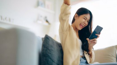 Happy young asian woman relax on comfortable couch at home texting messaging on smartphone, smiling girl use cellphone, chatting online message, shopping online from home