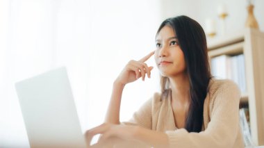 Bilgisayarda çalışan Asyalı genç bir kadın evdeki oturma odasında masa başında çalışıyor.