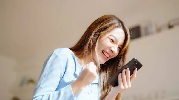 Funny euphoric young asian woman celebrating winning or getting ecommerce shopping offer on mobile smartphone. Excited happy girl winner looking smart phone celebrating success