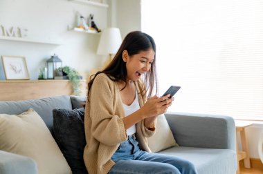 Mutlu genç Asyalı kadın rahat bir kanepede rahat rahat oturup akıllı telefonuna mesaj atıyor, gülümseyen kız cep telefonu sohbetini kullanıyor, cihazda kablosuz interneti tarıyor, evden online alışveriş yapıyor.