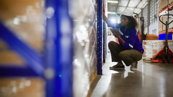 Young black woman warehouse workers holding digital tablet checking inventory management packaging boxes. Afro woman staff wearing vest and safety helmet and sitting count the box at storehouse