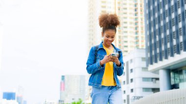 Zeki telefon kullanan ve şehirde yürürken gülümseyen genç siyahi bir kadın. Mutlu Afrikalı öğrenci, cep telefonuyla bir binanın önünden geçen genç bayan.