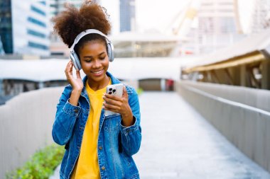 Cheerful black student girl wearing headset and using mobile smartphone, Walking at college building outdoor, Happy and smiling woman teenager. Modern education