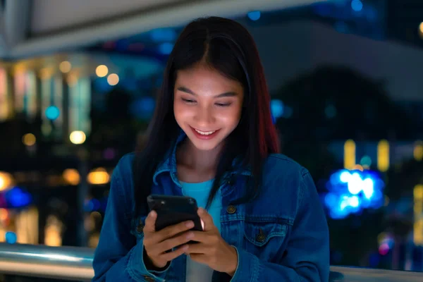 Beautiful young asian woman walking in city street at night using mobile smartphone. Happy woman standing and typing messages on her mobile phone