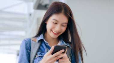 Dışarıda cep telefonu kullanan Asyalı genç bir kadın gülümsüyor. Kot ceket giyen ve halka açık bir yerde akıllı telefon tutan mutlu kadın turist.