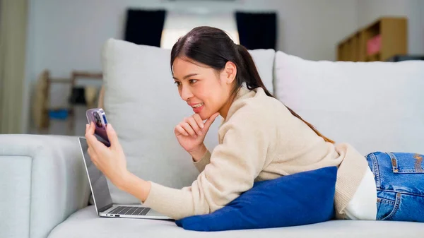 自宅でスマートフォンを使っている若いアジア人女性 ソファでリラックスしながら 女性が家でビデオ通話会議スマートフォンを作る笑顔 メッセージングやソーシャルネットワークを参照してください — ストック写真