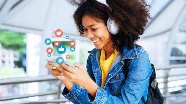 Genç siyahi kadınlar canlı yayın videosu izliyor ve kulaklık takıyor. Mutlu Afrikalı kadın eğlence için cep telefonu sosyal medya uygulaması kullanıyor