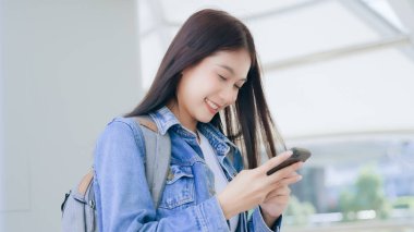 Dışarıda cep telefonu kullanan Asyalı genç bir kadın gülümsüyor. Kot ceket giyen ve halka açık bir yerde akıllı telefon tutan mutlu kadın turist.