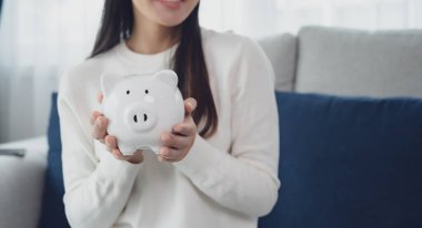 Genç Asyalı kadın beyaz domuz kumbarasını evde tutuyor. Para ve yatırım kavramını sakla