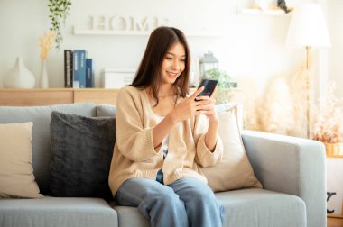 Happy young asian woman relax on comfortable couch at home texting messaging on smartphone, smiling girl use cellphone chatting, browse wireless internet on gadget, shopping online from home