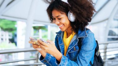 Açık havada sarı gömlek ve kot ceket giyerken kablosuz kulaklıkla müzik dinleyen mutlu Afro kadın.