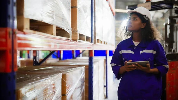 Young black woman warehouse workers holding digital tablet checking inventory management packaging boxes. Afro woman staff wearing vest and safety helmet and walking count the box at storehouse