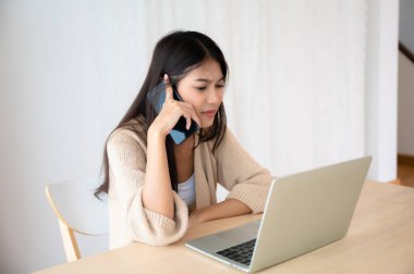 Evde bilgisayar bilgisayarı üzerinde çalışırken cep telefonuyla konuşan Asyalı genç bir kadın. Evde çalışmak, video konferansı, video görüşmesi.