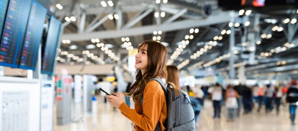 stock image Young asian woman in international airport, using mobile smartphone and checking flight at the flight information board