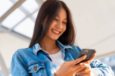 Dışarıda cep telefonu kullanan Asyalı genç bir kadın gülümsüyor. Kot ceket giyen ve halka açık bir yerde akıllı telefon tutan mutlu kadın turist.