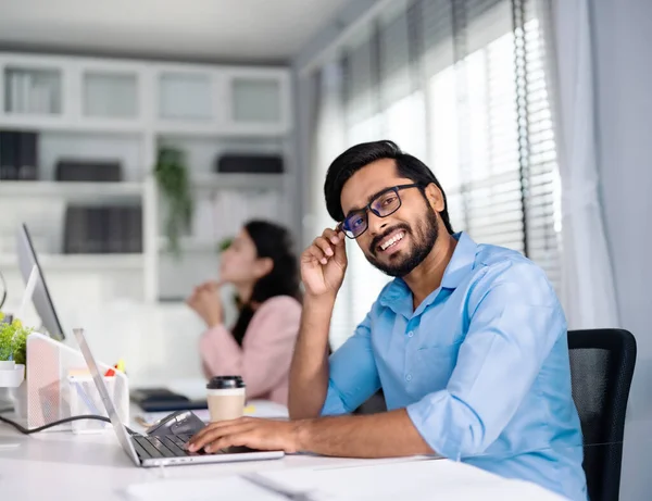 印度职业商人 戴着眼镜在电脑上工作的商人 直接看着相机 友善地微笑着 图库图片