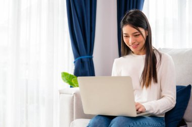 Genç Asyalı kadın evdeki koltukta otururken dizüstü bilgisayar kullanıyor.