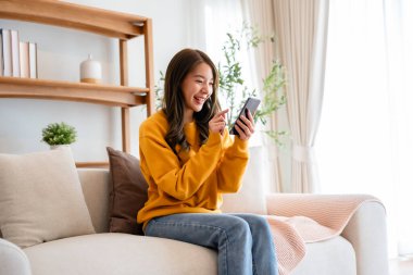 Happy young asian woman relax on comfortable couch at home texting messaging on smartphone, smiling girl use cellphone chatting, browse wireless internet on gadget, shopping online from home