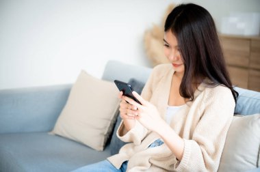 Happy young asian woman relax on comfortable couch at home texting messaging on smartphone, smiling girl use cellphone chatting, browse wireless internet on gadget, shopping online from home