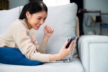 Happy young asian woman relax on comfortable couch at home texting messaging on smartphone, smiling girl use cellphone, browse wireless internet on gadget, shopping online from home