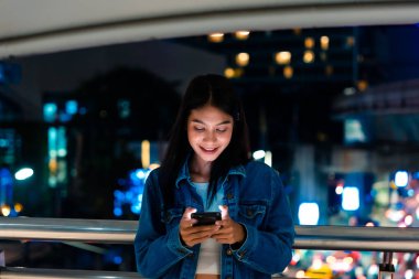 Güzel genç Asyalı kadın, neon ışıklarla dolu gece caddesinde akıllı telefon kullanıyor. Cep telefonu kullanan kadın