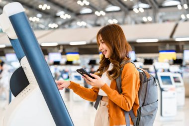 Havaalanı terminalinde kendini kontrol eden Asyalı genç bir kadın. Mutlu ve gülen kız internetten akıllı telefondan giriş yapıyor ve uluslararası havaalanından bilet alıyor.
