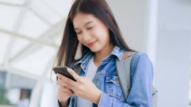 Dışarıda cep telefonu kullanan Asyalı genç bir kadın gülümsüyor. Kot ceket giyen ve halka açık bir yerde akıllı telefon tutan mutlu kadın turist.