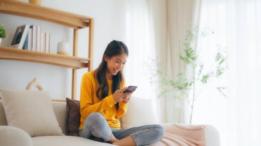 Mutlu genç Asyalı kadın evdeki rahat kanepede gevşiyor, akıllı telefondan sohbet mesajı yazıyor, gülümseyen kız cep telefonu sohbetini kullanıyor, tarayıcı kablosuz internetten bilgi arıyor, online alışveriş yapıyor.