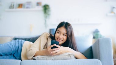 Happy young asian woman relax on comfortable couch at home texting messaging on smartphone, smiling girl use cellphone, browse wireless internet on gadget, shopping online from home