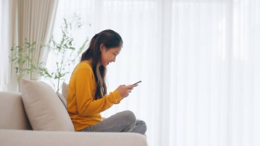 Mutlu genç Asyalı kadın evdeki rahat kanepede gevşiyor, akıllı telefondan sohbet mesajı yazıyor, gülümseyen kız cep telefonu sohbetini kullanıyor, tarayıcı kablosuz internetten bilgi arıyor, online alışveriş yapıyor.