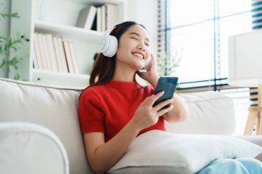 Genç Asyalı kadın evdeki kanepede dinleniyor, kulaklıkla cep telefonuyla müzik dinliyor. Halk yaşam tarzı konsepti