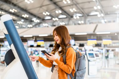 Havaalanı terminalinde kendini kontrol eden Asyalı genç bir kadın. Mutlu ve gülen kız internetten akıllı telefondan giriş yapıyor ve uluslararası havaalanından bilet alıyor.