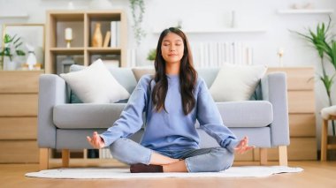 Mutlu genç Asyalı kadın evde yoga ve meditasyon yapıyor oturma odasında lotus pozisyonunda oturuyor ve gözleri kapalı dinleniyor. Dikkatli meditasyon ve refah kavramı