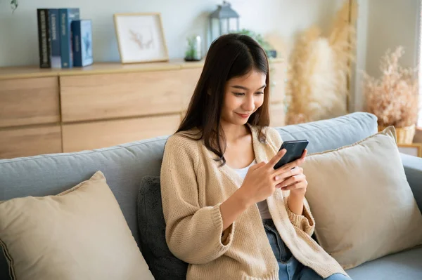 Happy young asian woman relax on comfortable couch at home texting messaging on smartphone, smiling girl use cellphone chatting, browse wireless internet on gadget, shopping online from home