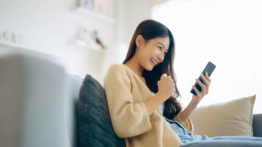 Happy young asian woman relax on comfortable couch at home texting messaging on smartphone, smiling girl use cellphone, chatting online message, shopping online from home