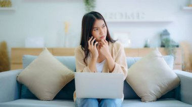 Genç Asyalı kadın evdeki koltukta otururken bilgisayar ve cep telefonu kullanıyor.