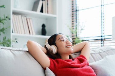 Rahat genç Asyalı kadın rahat koltukta dinleniyor, sakin çekici kız kanepede dinleniyor, temiz hava soluyor gözleri kapalı, gözlük takıyor ve kulaklık takıyor, evde meditasyon yapıyor.