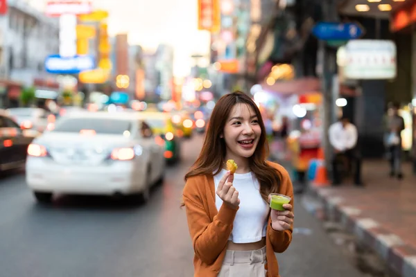 stock image Young Asian tourists standing and eating Thai desserts. Young beautiful tourists in Chinatown street food market, Bangkok, Thailand