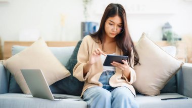 Genç Asyalı kadın gözlük takıyor ve evdeki koltukta otururken tablet kullanıyor.