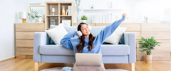 Young asian woman in good spirits working on laptop at home while sitting on the floor close to the couch. Excited female using computer notebook, wearing headphones and celebrating success