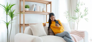 Cep telefonu teknolojisiyle kanepede oturan mutlu genç Asyalı kadın ecommerce alışverişi yapıyor, internetten alışveriş yapıyor evdeki rahat oturma odasındaki kanepede rahatlatıcı mesajlar yazıyor.