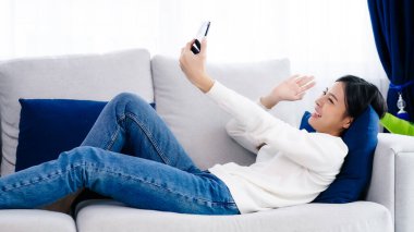 Happy young asian woman relaxing at home. Female smile lying on sofa and holding mobile smartphone. Girl using video call to friend